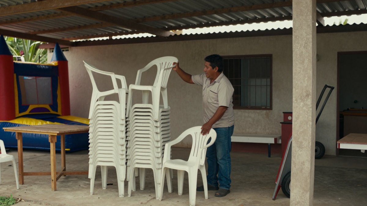 Hombre mexicano acomoda pilas de sillas de plástico blancas en un patio techado con brincolín al fondo